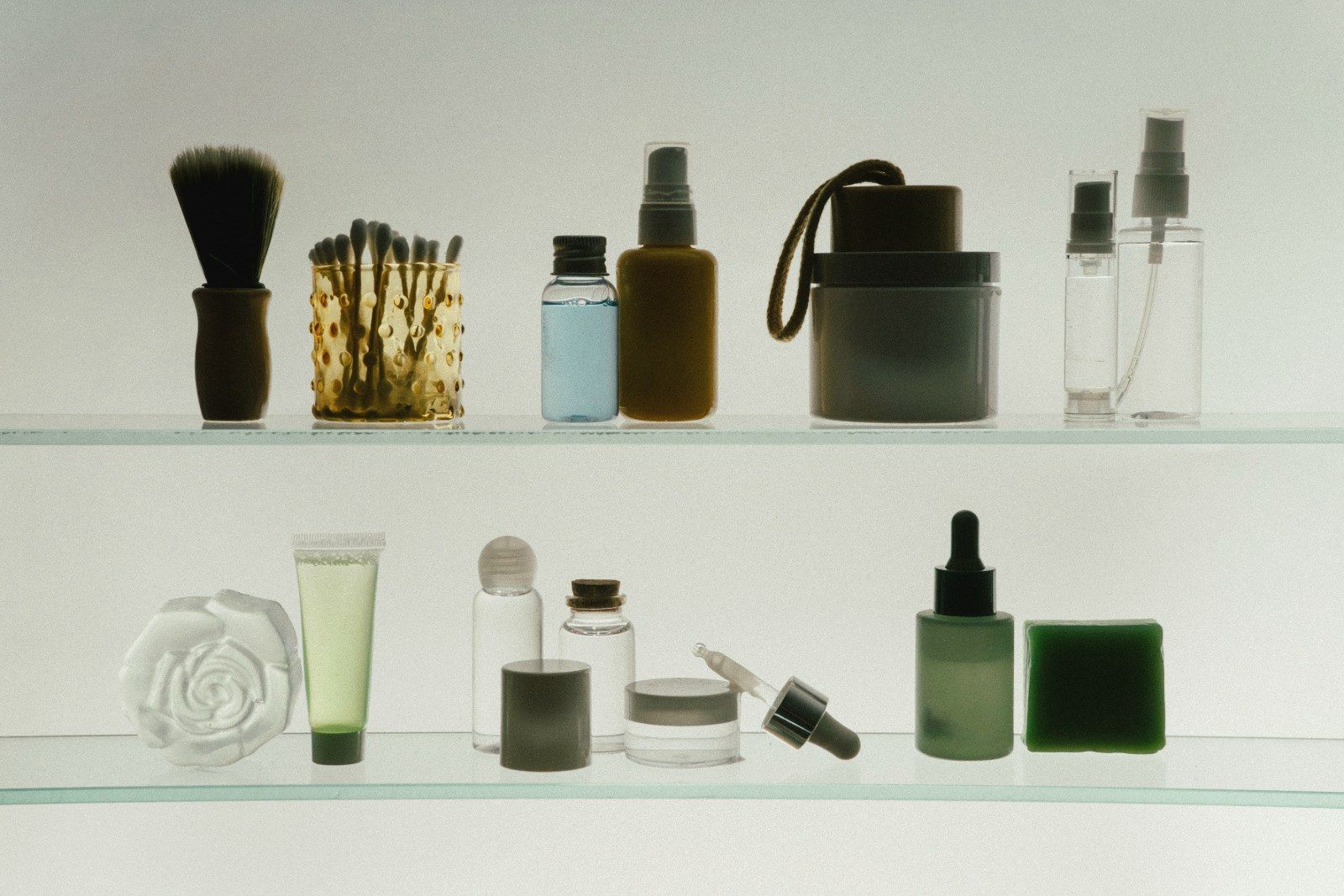 Cosmetic bottles, jars and a brush arranged on two glass shelves.