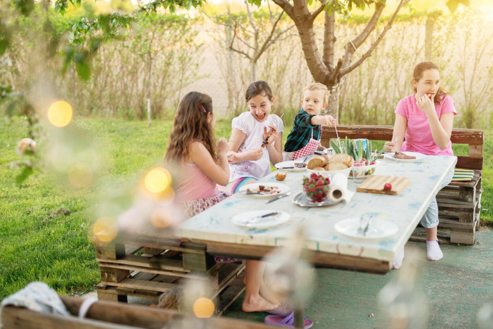 Four children eating at a pallet table in a sunlit garden.