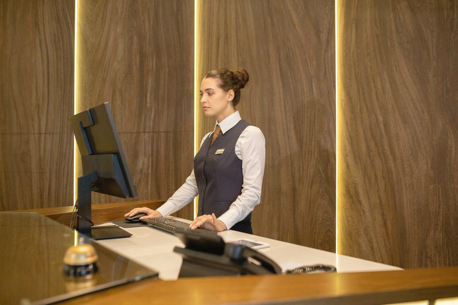A receptionist working at a computer behind a wood-panelled hotel desk with a bell on the counter.