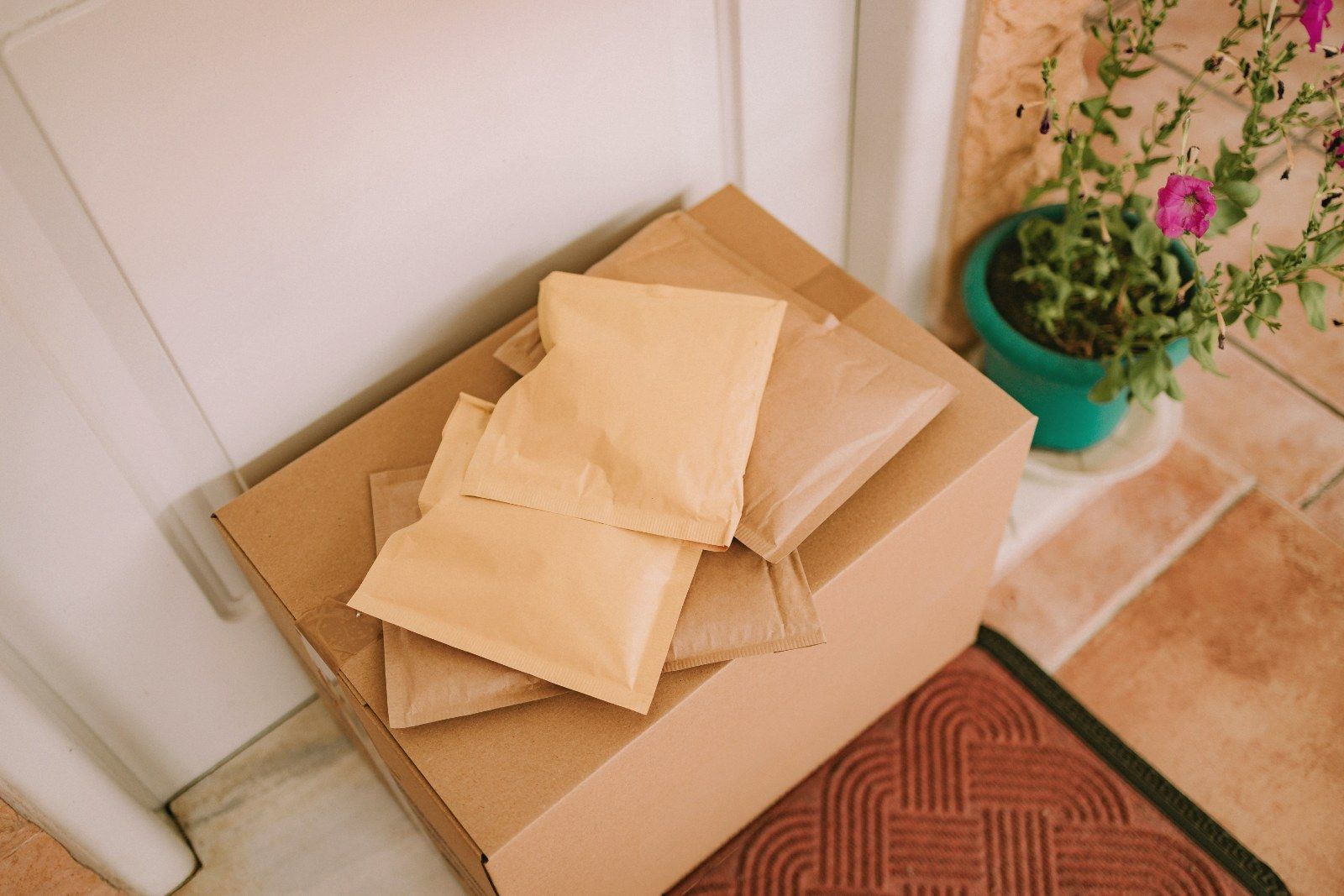 A stack of parcels and padded envelopes on a doorstep beside a potted plant and a doormat.