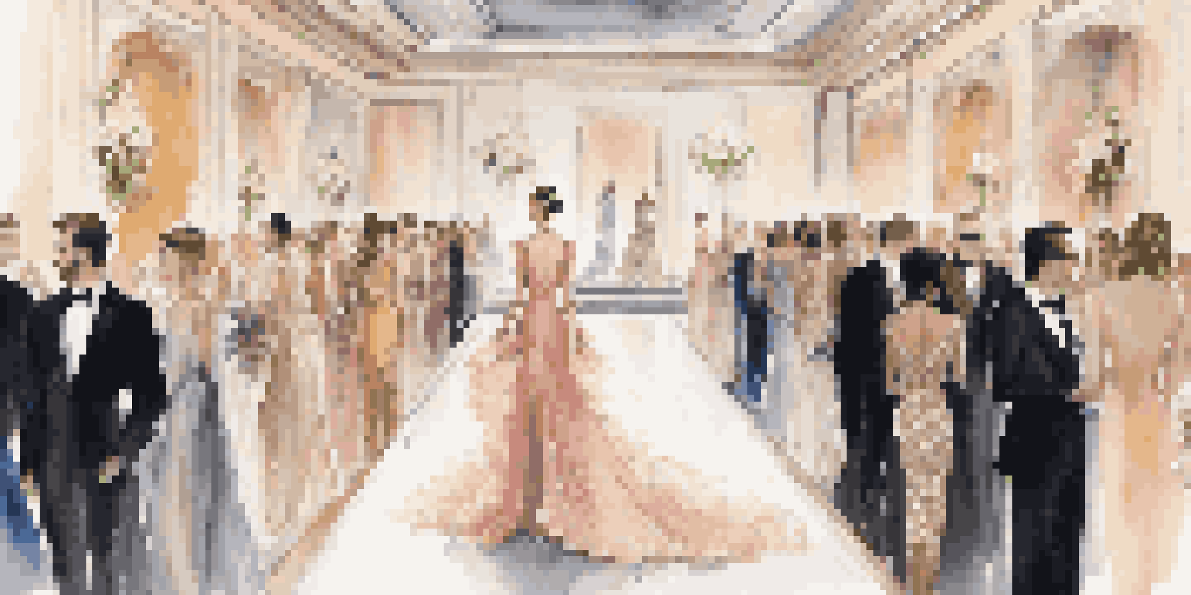 A fashion show with models on a runway wearing elegant garments, surrounded by an audience in formal attire and decorated with floral arrangements.