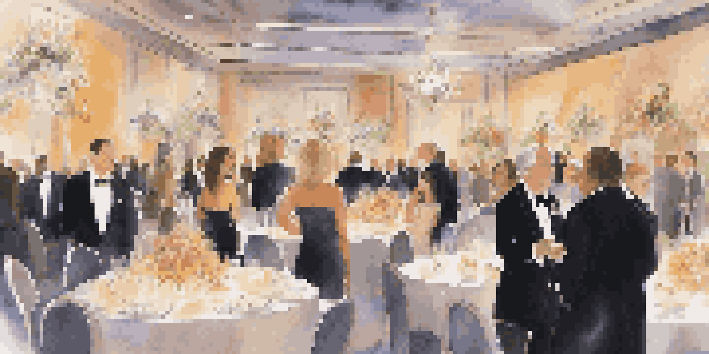 A fancy charity event with guests in formal attire, surrounded by flowers and candles, while a video plays in the background.