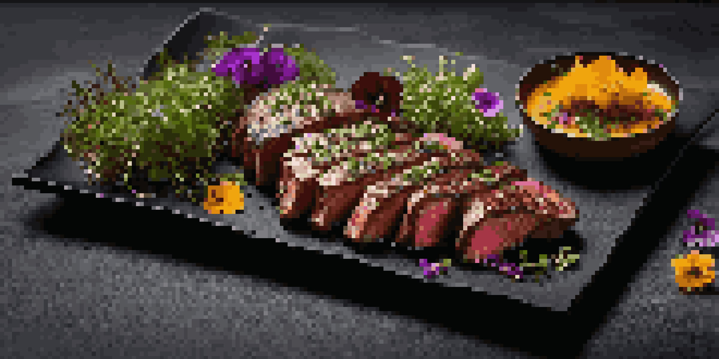 A gourmet dish of sous-vide steak with herb sauce, microgreens, and edible flowers on a dark slate plate, illuminated by warm lighting.