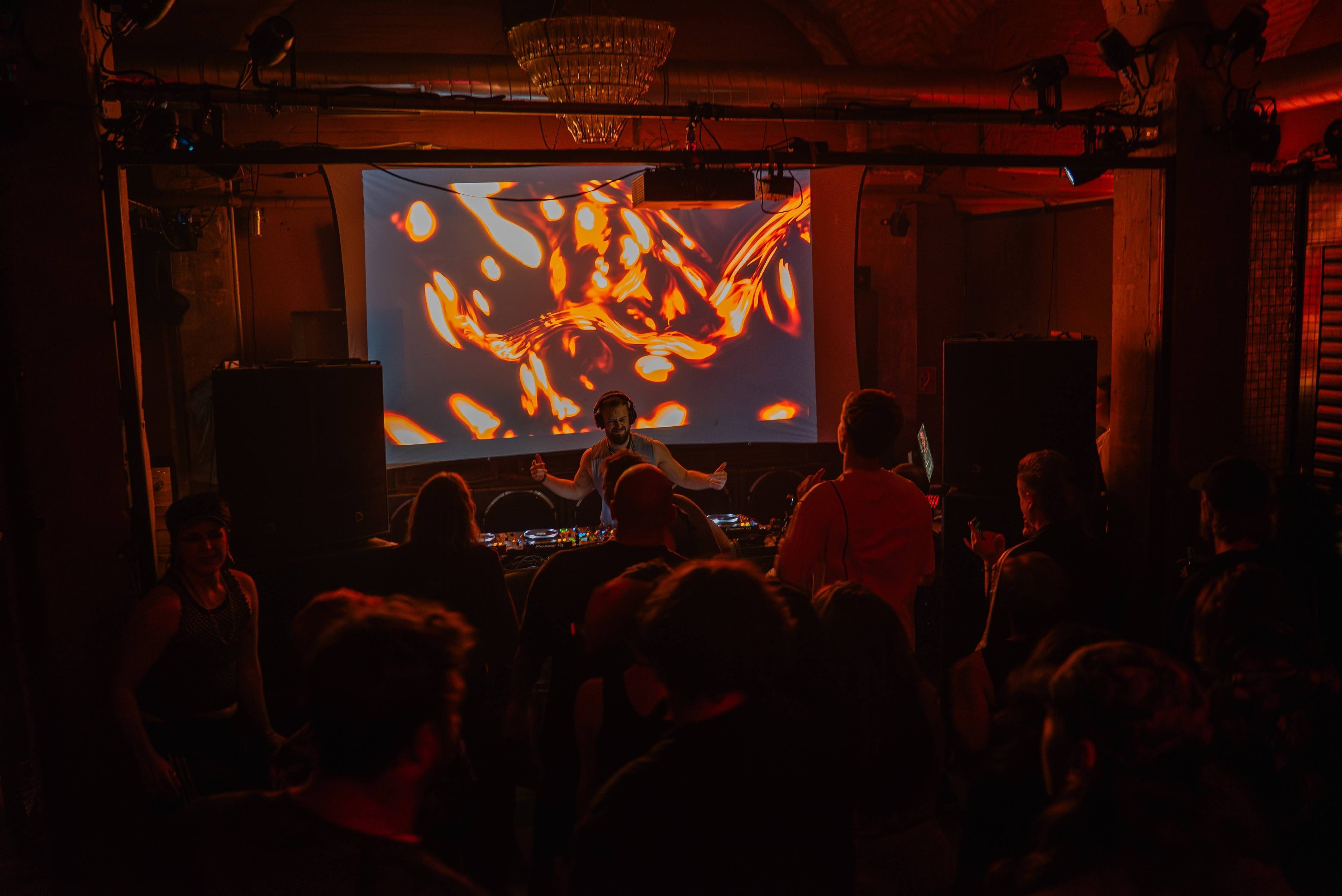 SKVLLFACE performing to a lively crowd in a dimly lit club, with vibrant abstract visuals projected on a screen behind.