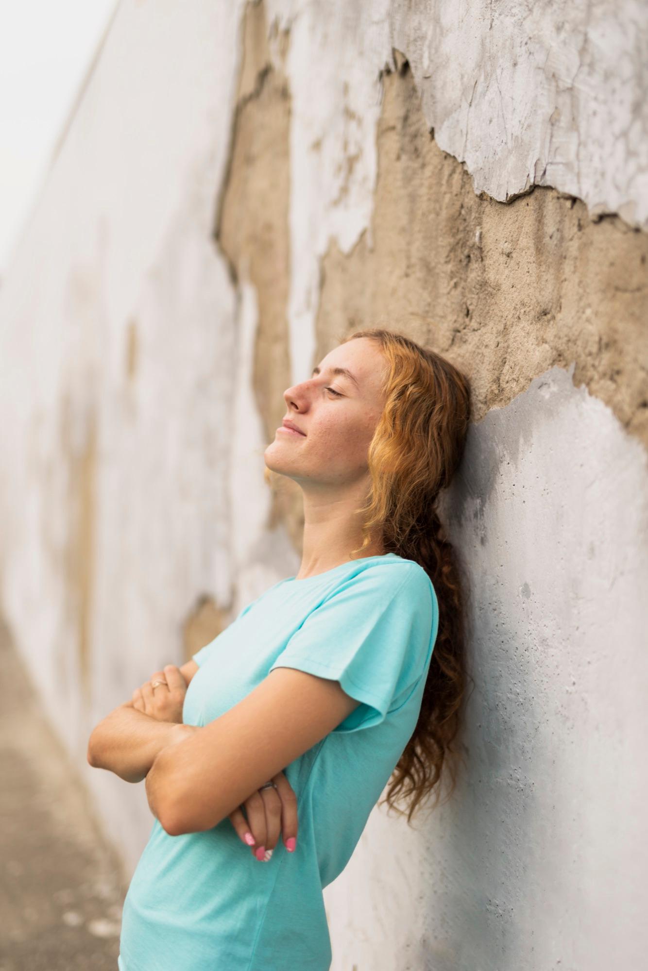 A young person leaning against a wall with eyes closed, smiling in the sun