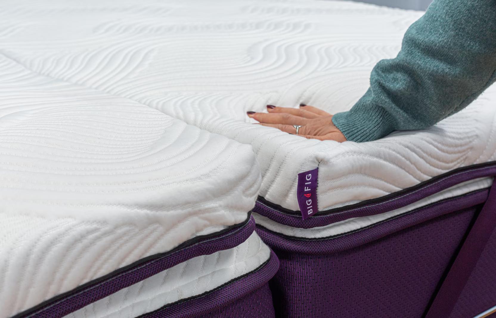 Hand pressing into the Big Fig Micro-Coil Topper, a white. quilted surface bedding accessory on a deep fig colored Big Fig mattress. The topper's corner tag is visible, labeled "BIG FIG".