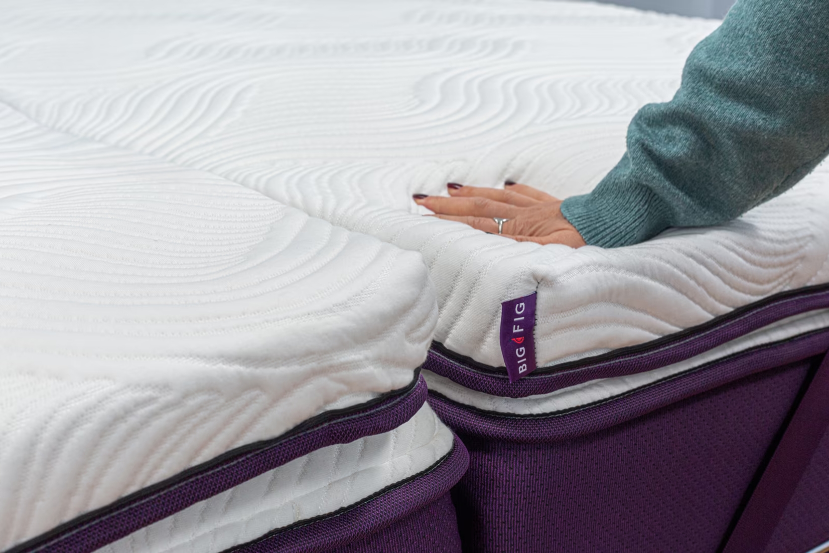 Hand pressing into the Big Fig Micro-Coil Topper, a white. quilted surface bedding accessory on a deep fig colored Big Fig mattress. The topper's corner tag is visible, labeled "BIG FIG".