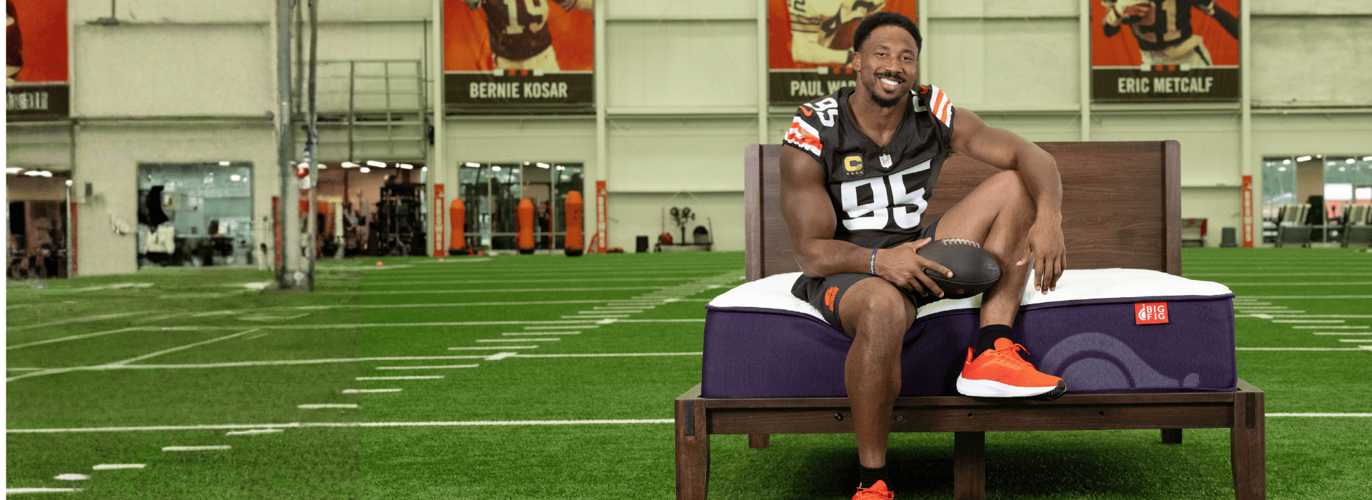 Myles Garrett in his Browns' #95 jersey sitting on a Big Fig Mattress with a white top and deep fig base in the Browns' indoor practice field with banners of ex-Browns legends hanging in the background.