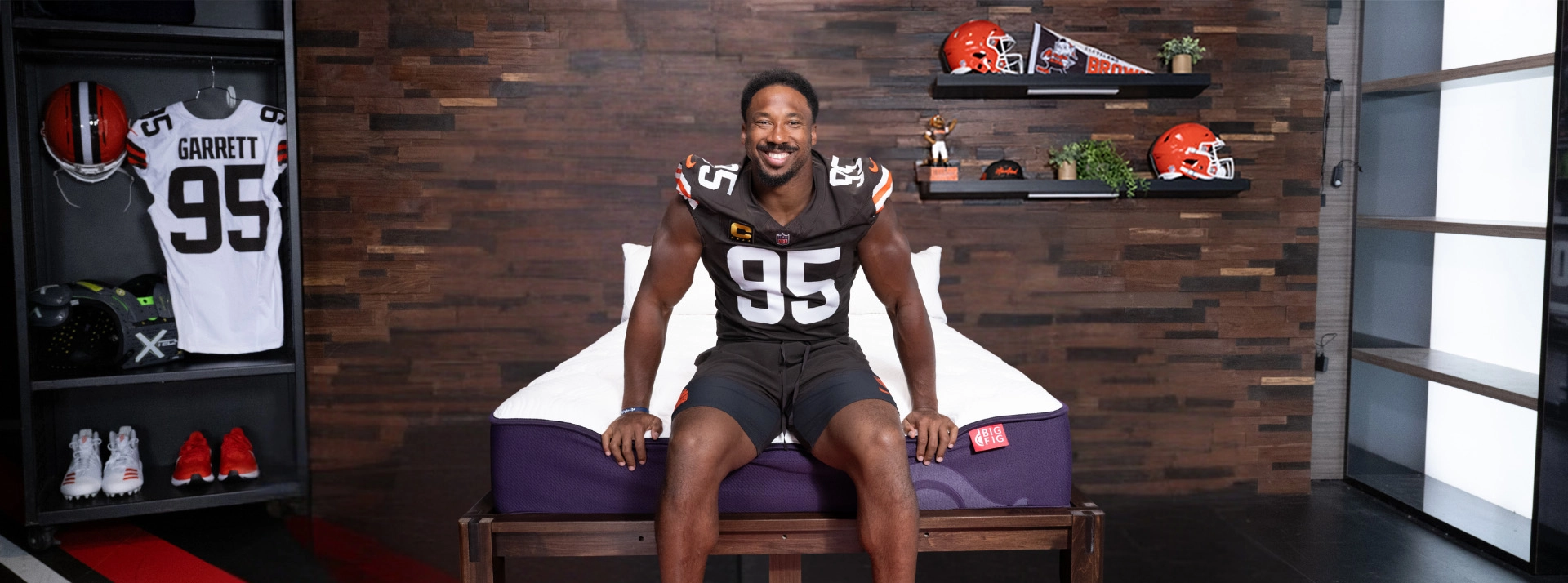 Myles Garrett in his Cleveland Browns #95 jersey sitting on a Big Fig Mattress: white top, deep fig colored base, in front of a dark wood wall decorated with Browns' helmets and team gear.