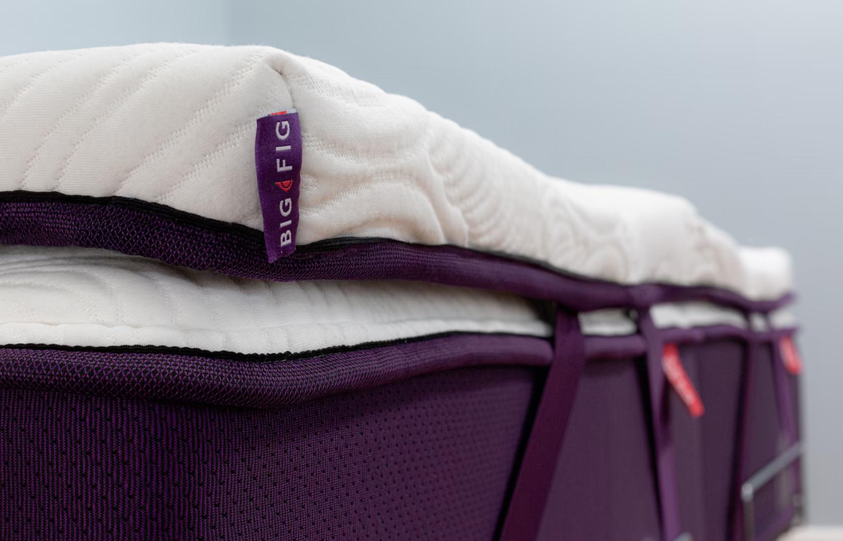 Big Fig Micro-Coil Topper with white quilted wave surface secured to a deep fig Big Fig mattress using adjustable corner straps. Corner tag reads "Big Fig".