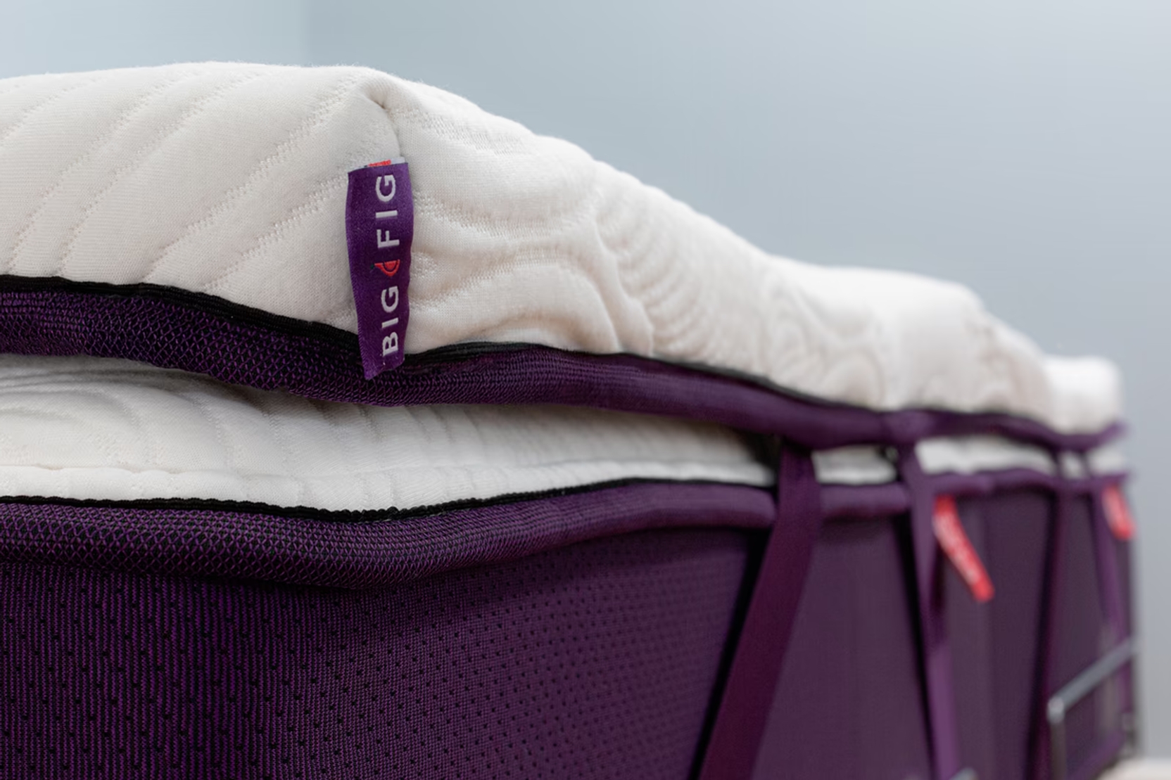 Big Fig Micro-Coil Topper with white quilted wave surface secured to a deep fig Big Fig mattress using adjustable corner straps. Corner tag reads "Big Fig".