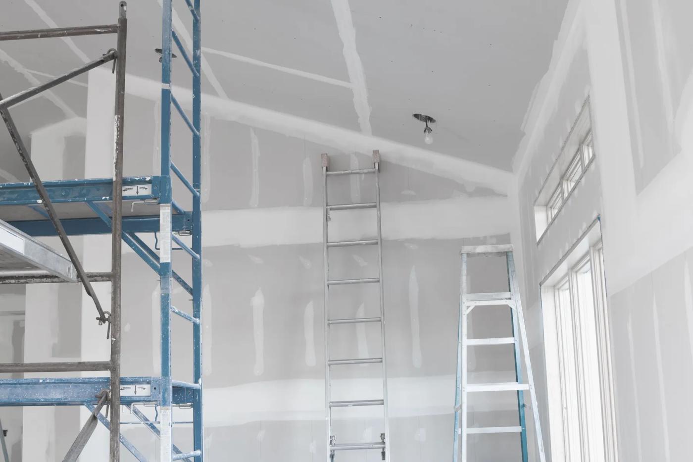 Unfinished home interior with taped drywall, scaffolding, and a ladder beside tall windows.