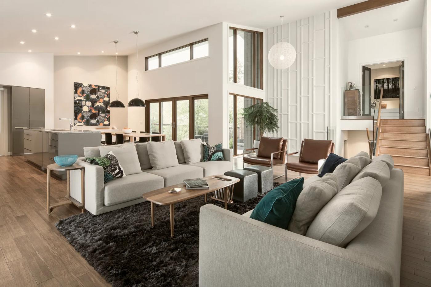 Double-height living room with neutral sofas, tall windows, and an adjoining open kitchen.