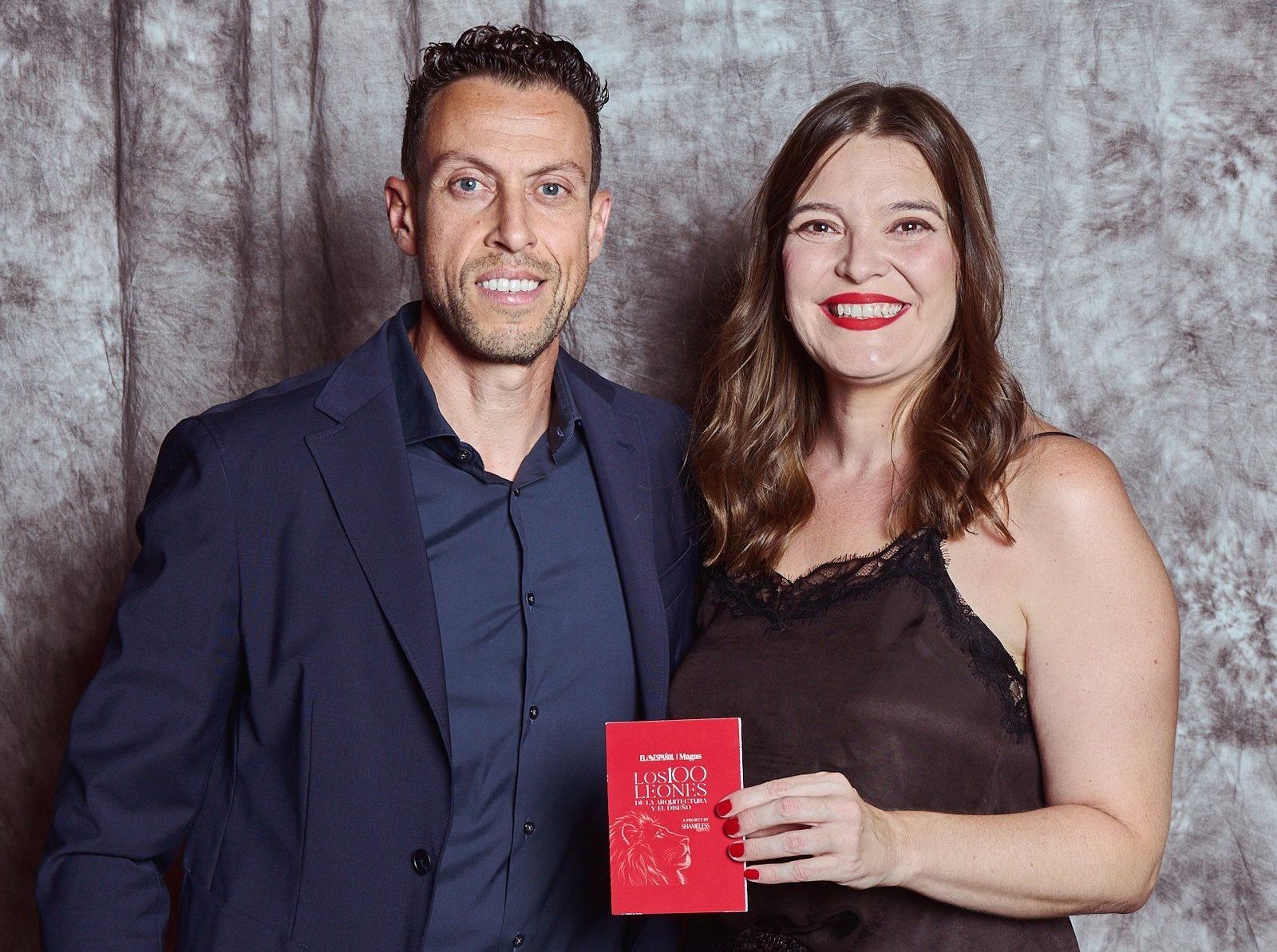 Portrait of a man and a woman smiling, she holds a red book, against a grey textile background.