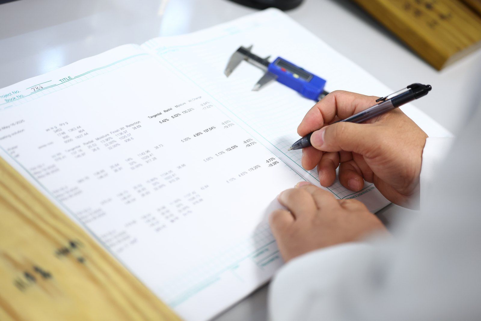 Hands recording measurements beside calipers and a wood sample