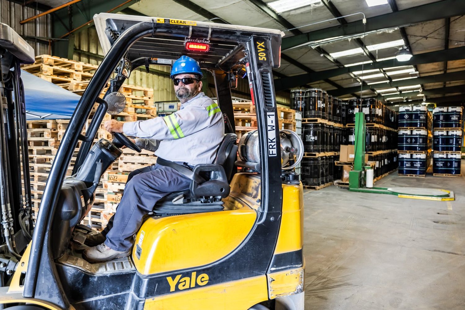 Forklift moving pallets past stacked drums in a warehouse