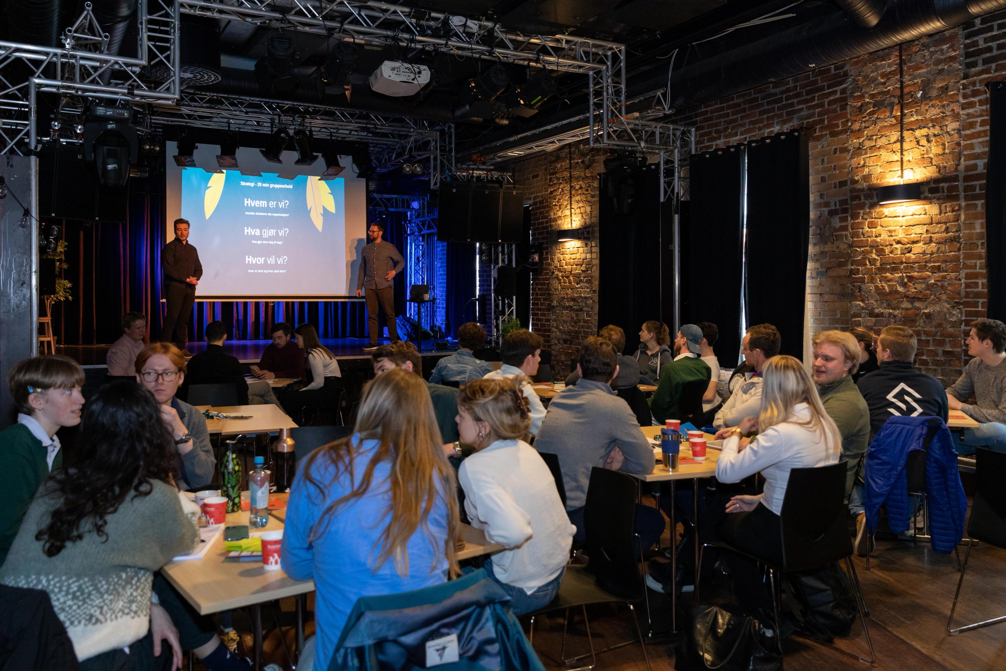 En gruppe unge voksne sitter ved bord i et mørkt lokale og følger med på en presentasjon på en scene, der to foredragsholdere står foran en stor skjerm med norsk tekst. Stemningen virker uformell og sosial, med drikke og notater på bordene.