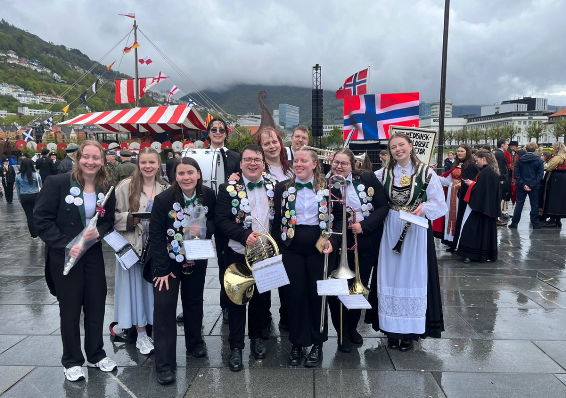 En glad gjeng unge musikere i uniform og bunad står samlet på en våt plass under 17. mai-feiring, med instrumenter, mange buttons og norske flagg i bakgrunnen.
