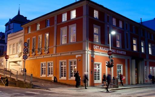 Kveldsbilde av den opplyste bygningen Det Akademiske Kvarter i Bergen, med mennesker som går forbi på fortauet foran.
