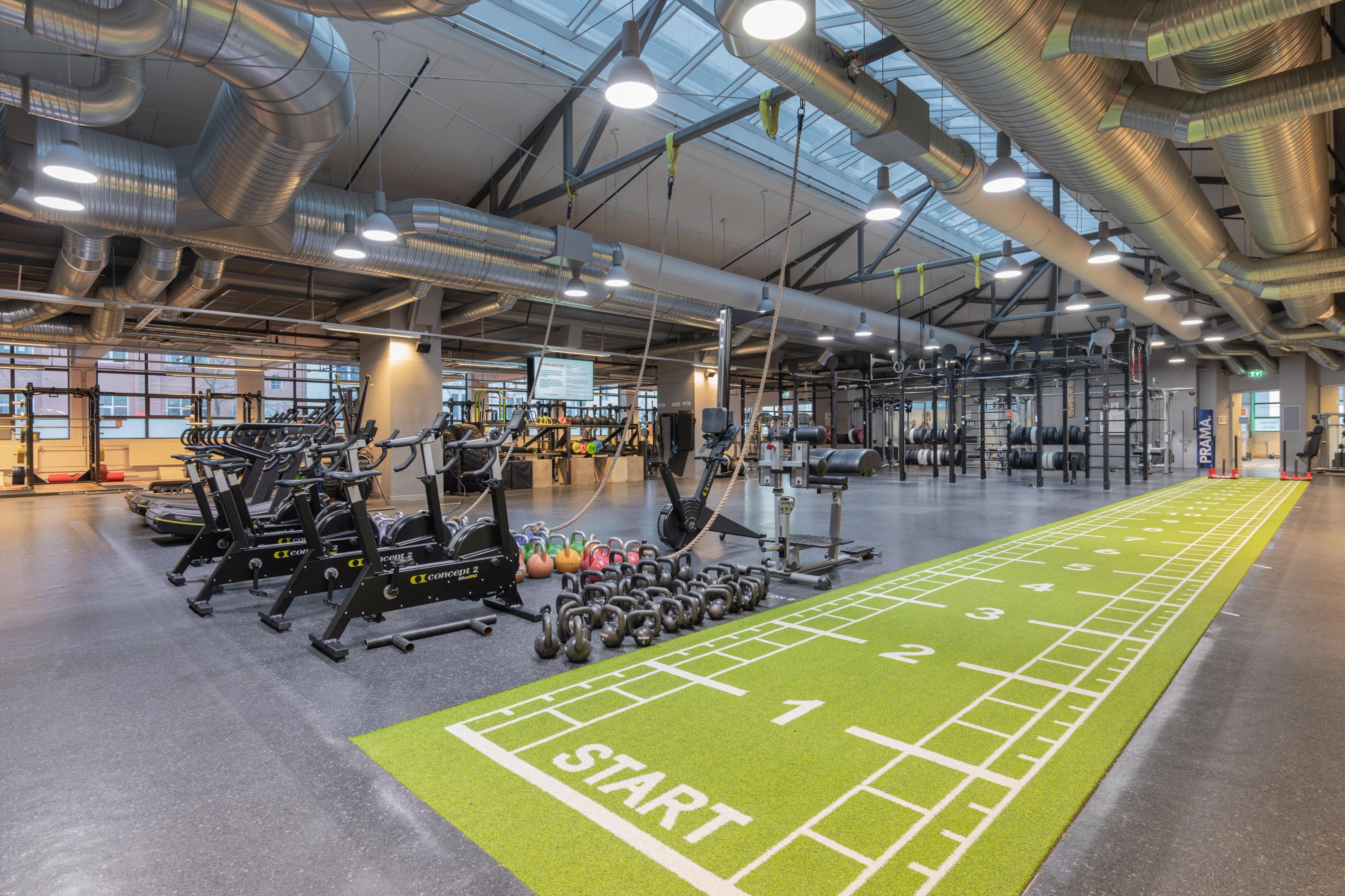 Stort, moderne treningssenter med kondisjonsapparater, frivekter og kettlebells ved siden av en grønn sprintbane merket med «START» og nummererte felt.