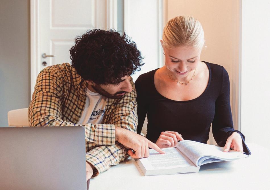 Two students are sitting at a table reading an open textbook, while one of them points at the text and a laptop is placed in front of them.