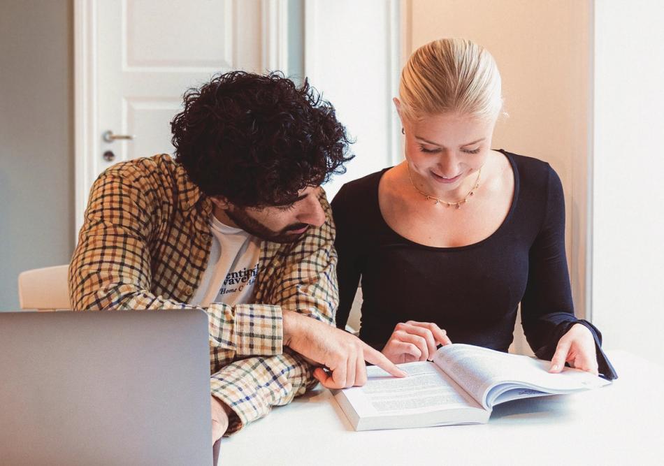 Two students are sitting at a table reading an open textbook, while one of them points at the text and a laptop is placed in front of them.