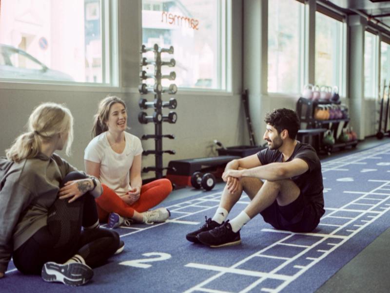 Tre personer sitter på gulvet i et treningssenter og snakker sammen etter en treningsøkt. Treningsutstyr som manualer og kettlebells står i bakgrunnen.