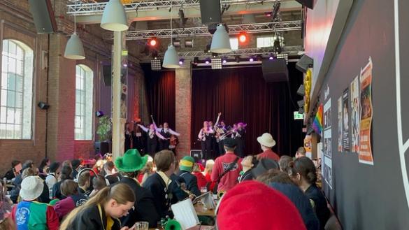 Et livlig publikum med fargerike hatter ser på en musikkopptreden på en opplyst scene i en stor industripreget sal.