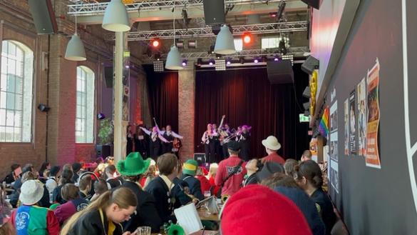 Et livlig publikum med fargerike hatter ser på en musikkopptreden på en opplyst scene i en stor industripreget sal.