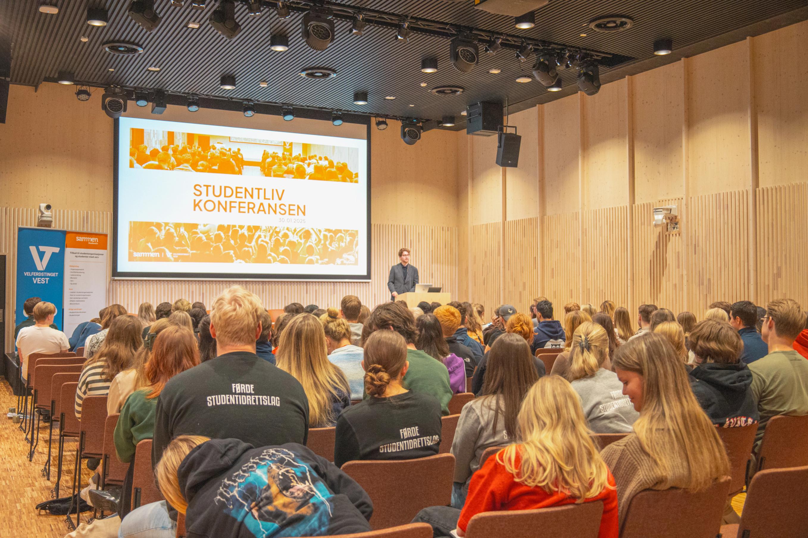A speaker stands in front of a large group of students in an auditorium during the “Student Life Conference”, with the presentation shown on a large screen behind him.