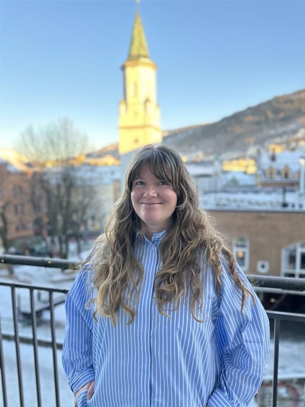 En person med langt, bølgete hår og blåstripet skjorte (Louise) står og smiler på en balkong, med en kirketårn og snødekte bygninger i bakgrunnen.