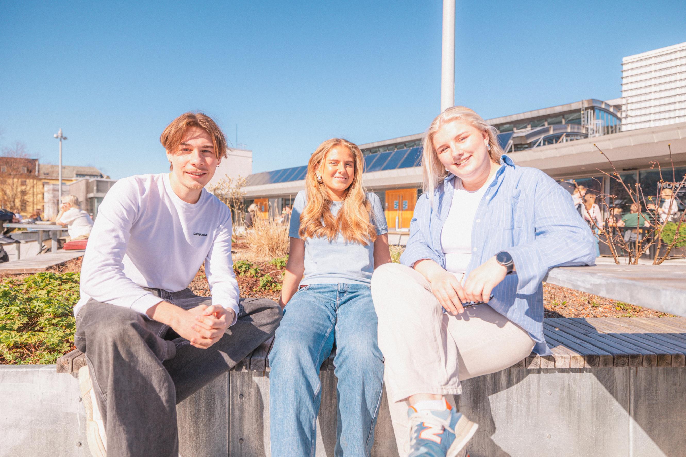 Tre unge personer sitter ute i sola på et campusområde og smiler mot kameraet, med moderne bygninger i bakgrunnen under blå himmel.