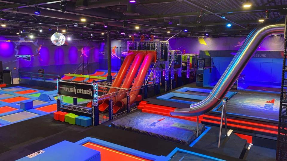 Innendørs aktivitetspark med trampoliner, hinderløyper og flere store sklier, opplyst av fargerike lys.