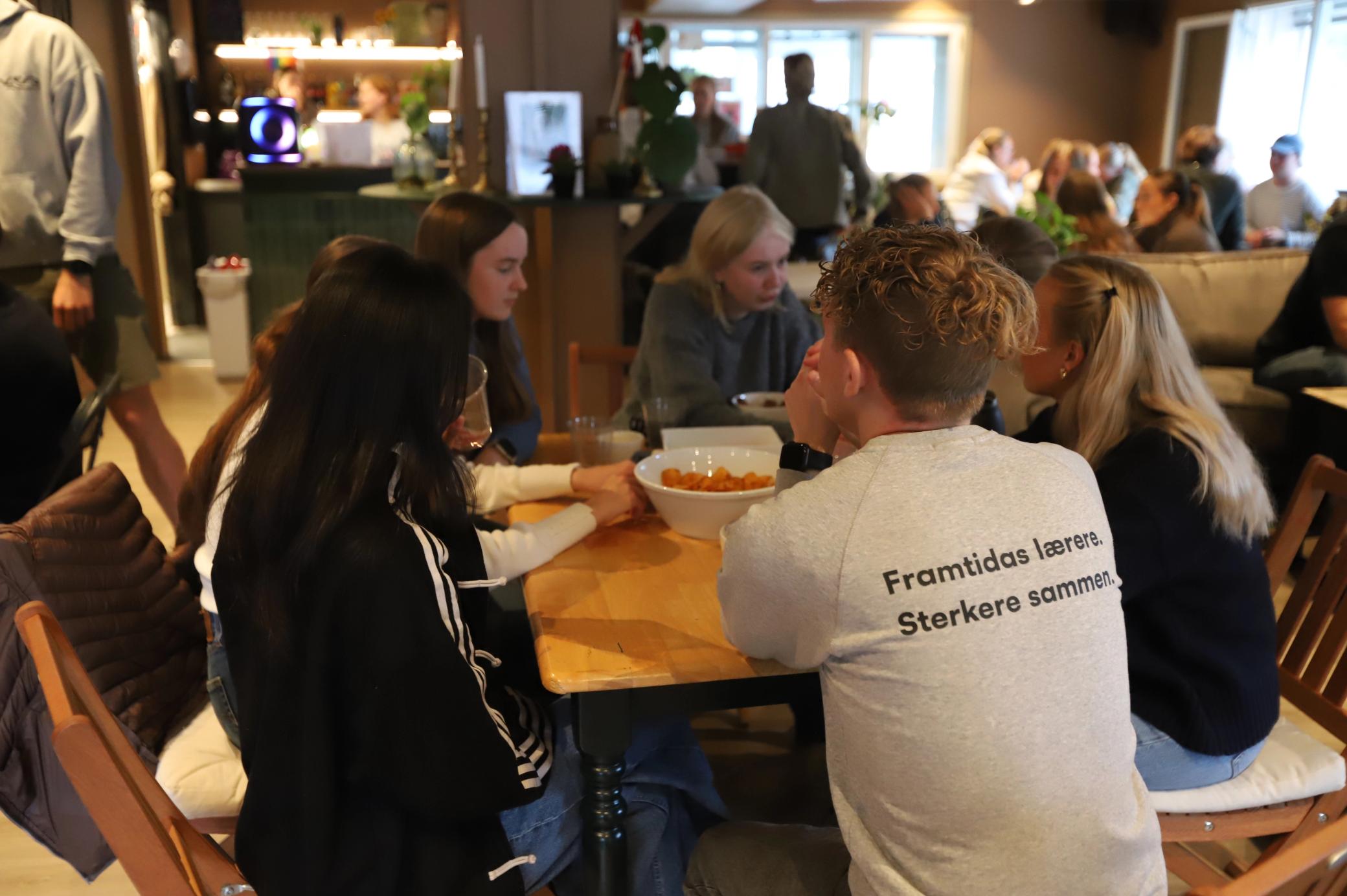 En gruppe unge mennesker sitter rundt et bord og prater i en travel kafé. På ryggen til en av dem står teksten «Framtidas lærere. Sterkere sammen.»