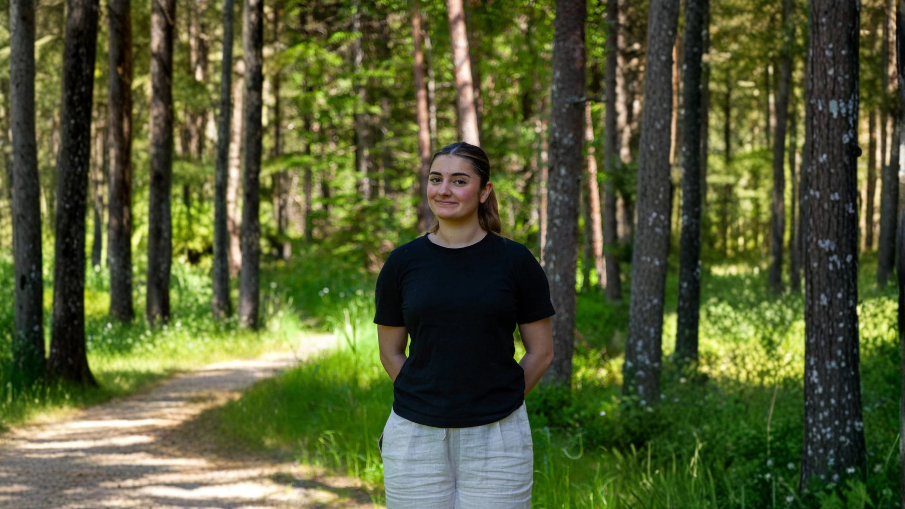 En smilende ung kvinne i svart T-skjorte og lyse bukser står på en solfylt sti i en grønn skog.