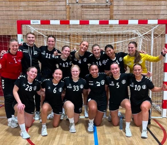 Et håndballag med smilende kvinnelige spillere i svarte drakter poserer samlet foran målet i en idrettshall.