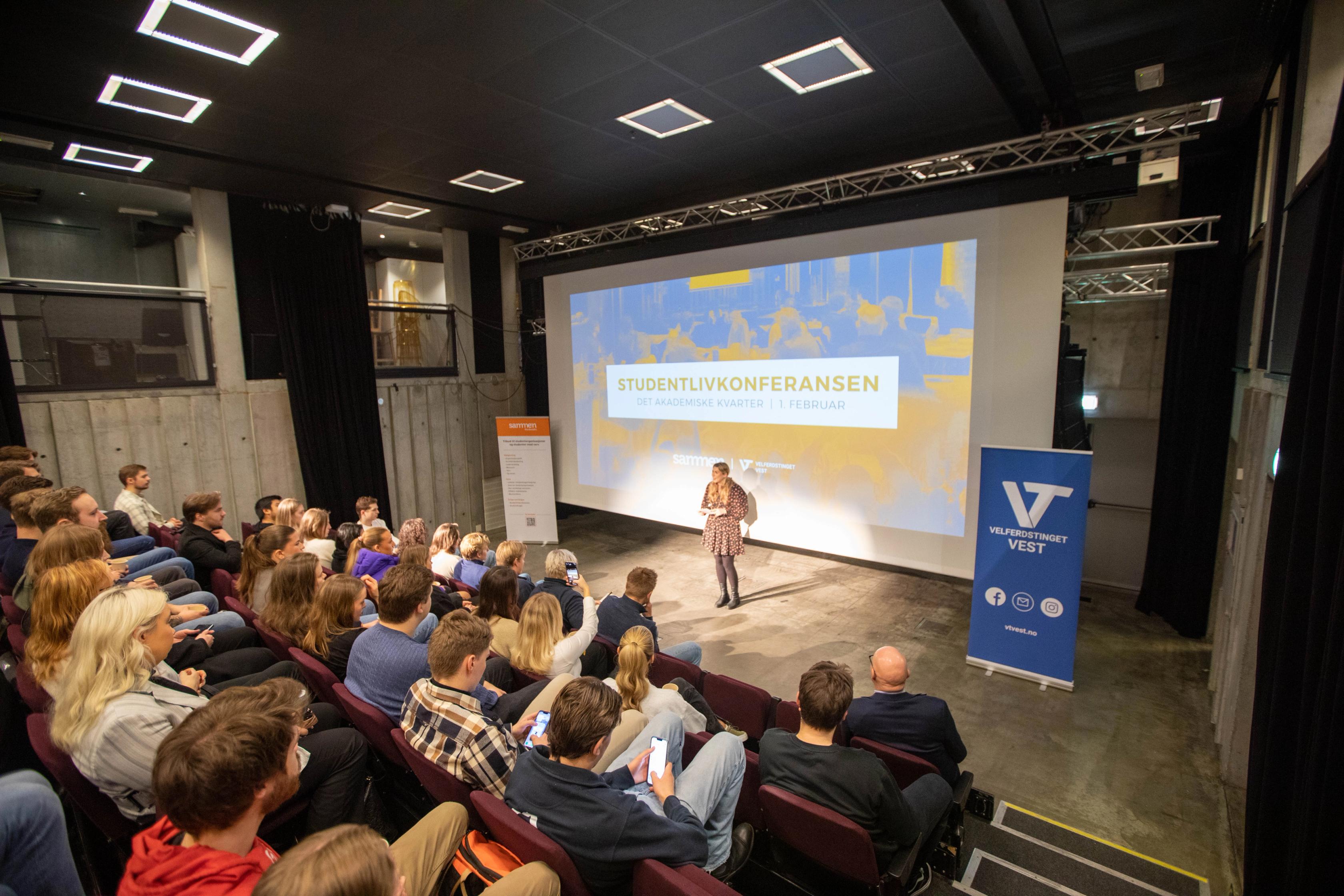En foredragsholder står på en scene foran en stor skjerm med teksten «Studentlivkonferansen», mens en sal full av studenter følger med fra stolradene. Til høyre står en blå roll-up med logoen til Velferdstinget Vest.