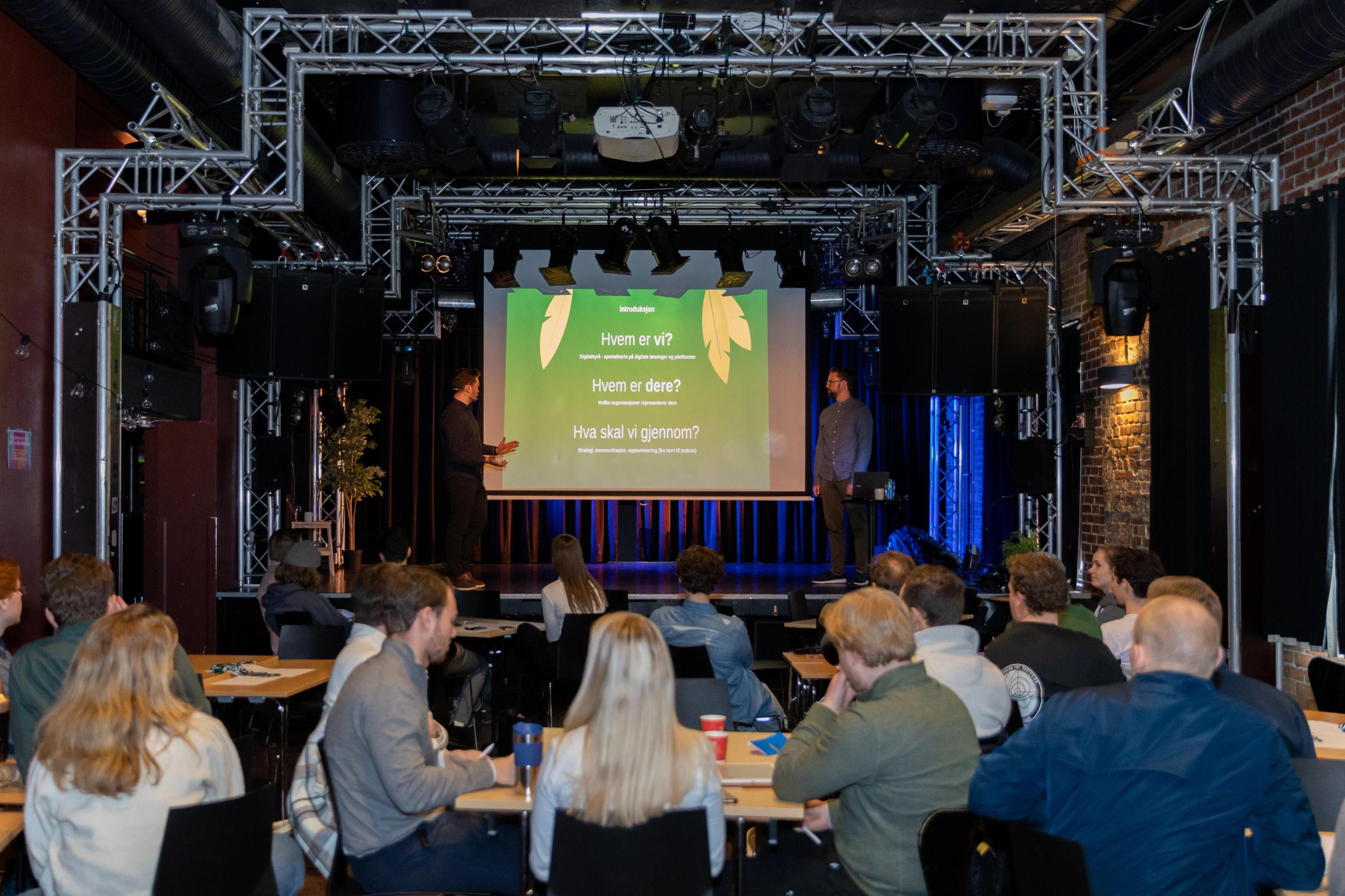 En gruppe mennesker sitter ved bord i en sal og følger med på en presentasjon, der to foredragsholdere står på en scene foran en stor skjerm med grønn bakgrunn og teksten «Hvem er vi? Hvem er dere? Hva skal vi gjennom?».