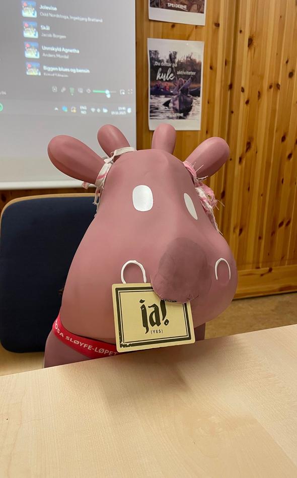 En rosa, tegneserieaktig elgfigur sitter ved et bord og holder et skilt i munnen der det står «ja! (yes)», med en projektorskjerm og plakater i bakgrunnen.