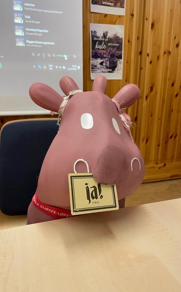 En rosa, tegneserieaktig elgfigur sitter ved et bord og holder et skilt i munnen der det står «ja! (yes)», med en projektorskjerm og plakater i bakgrunnen.