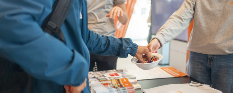 Close-up of people at an information table as one person reaches into a bowl of wrapped candy, with brochures and promotional materials spread out on the table.