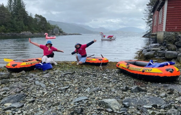 To personer i rosa T-skjorter poserer glade på en steinete strand ved en fjord, omgitt av oransje oppblåsbare båter, mens en oppblåsbar flamingo og enhjørning flyter ute på vannet foran fjell og skog.