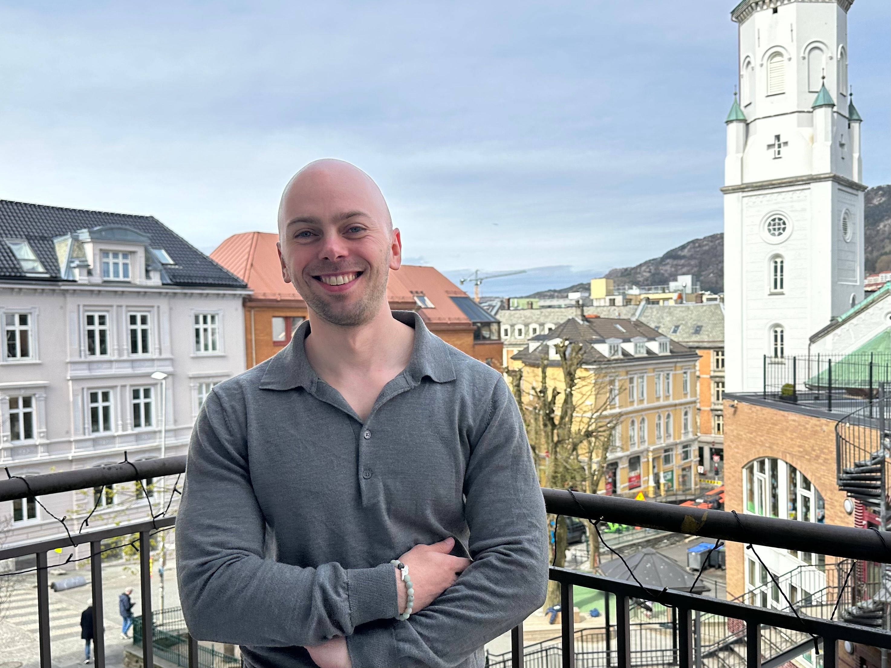 Smilende person (Henrik) står på en balkong med utsikt over en by med fargerike bygårder, en hvit kirketårn og fjell i bakgrunnen under en lys himmel.