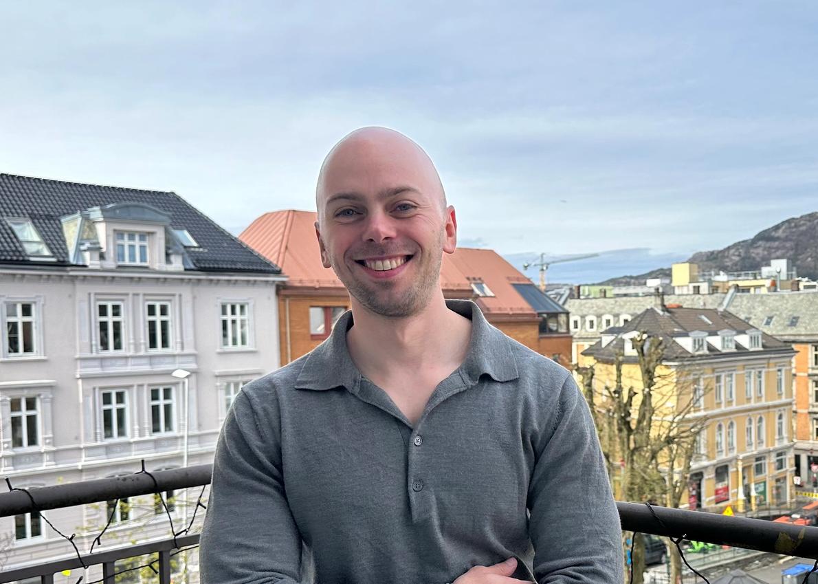 Smilende person (Henrik) står på en balkong med utsikt over en by med fargerike bygårder, en hvit kirketårn og fjell i bakgrunnen under en lys himmel.