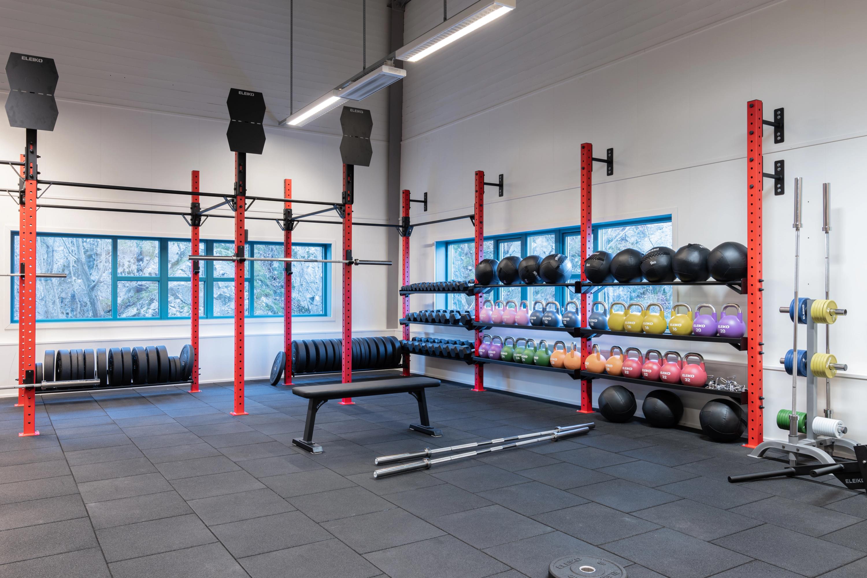 Et lyst og moderne treningsrom med røde styrkestativer, benk, vektstenger og hyller med fargerike kettlebells, manualer og medisinballer.