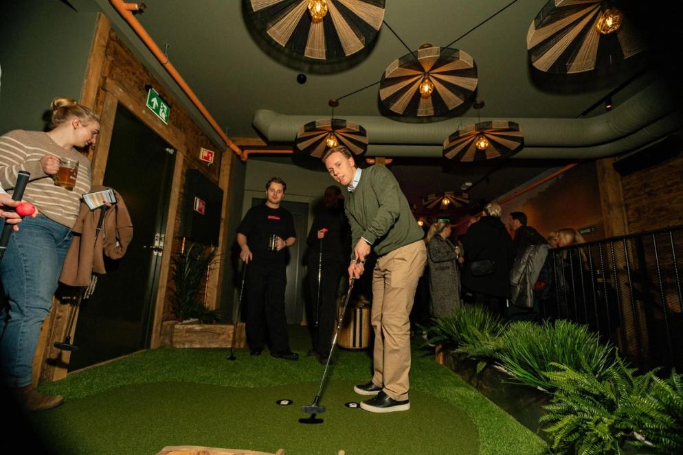 En mann spiller minigolf innendørs på en kunstgressbane, mens andre gjester står rundt med drikke og følger med i det stemningsfulle lokalet.