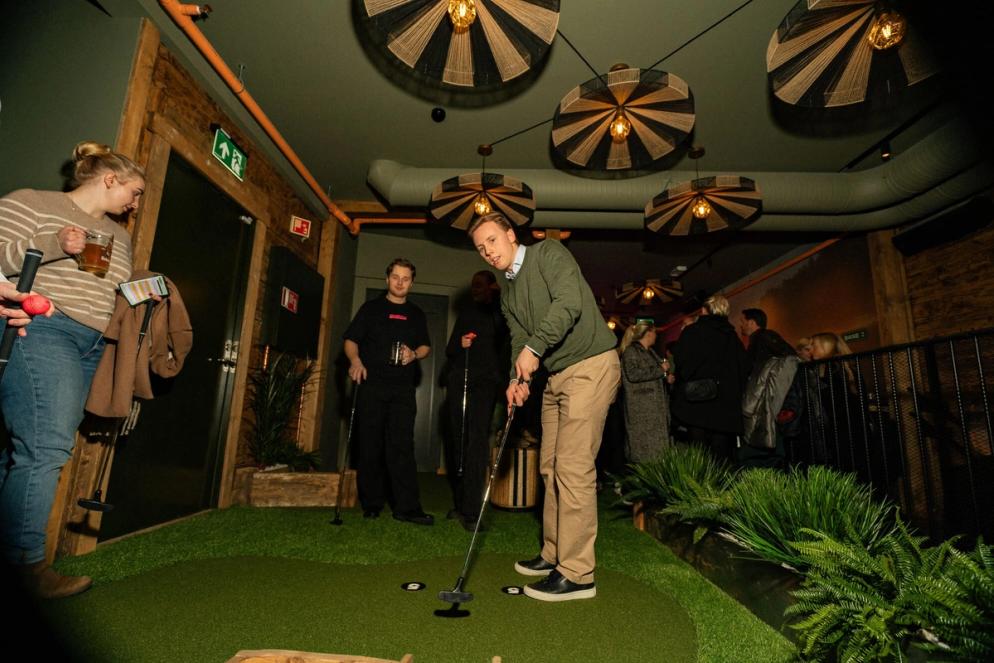 En mann spiller minigolf innendørs på en kunstgressbane, mens andre gjester står rundt med drikke og følger med i det stemningsfulle lokalet.