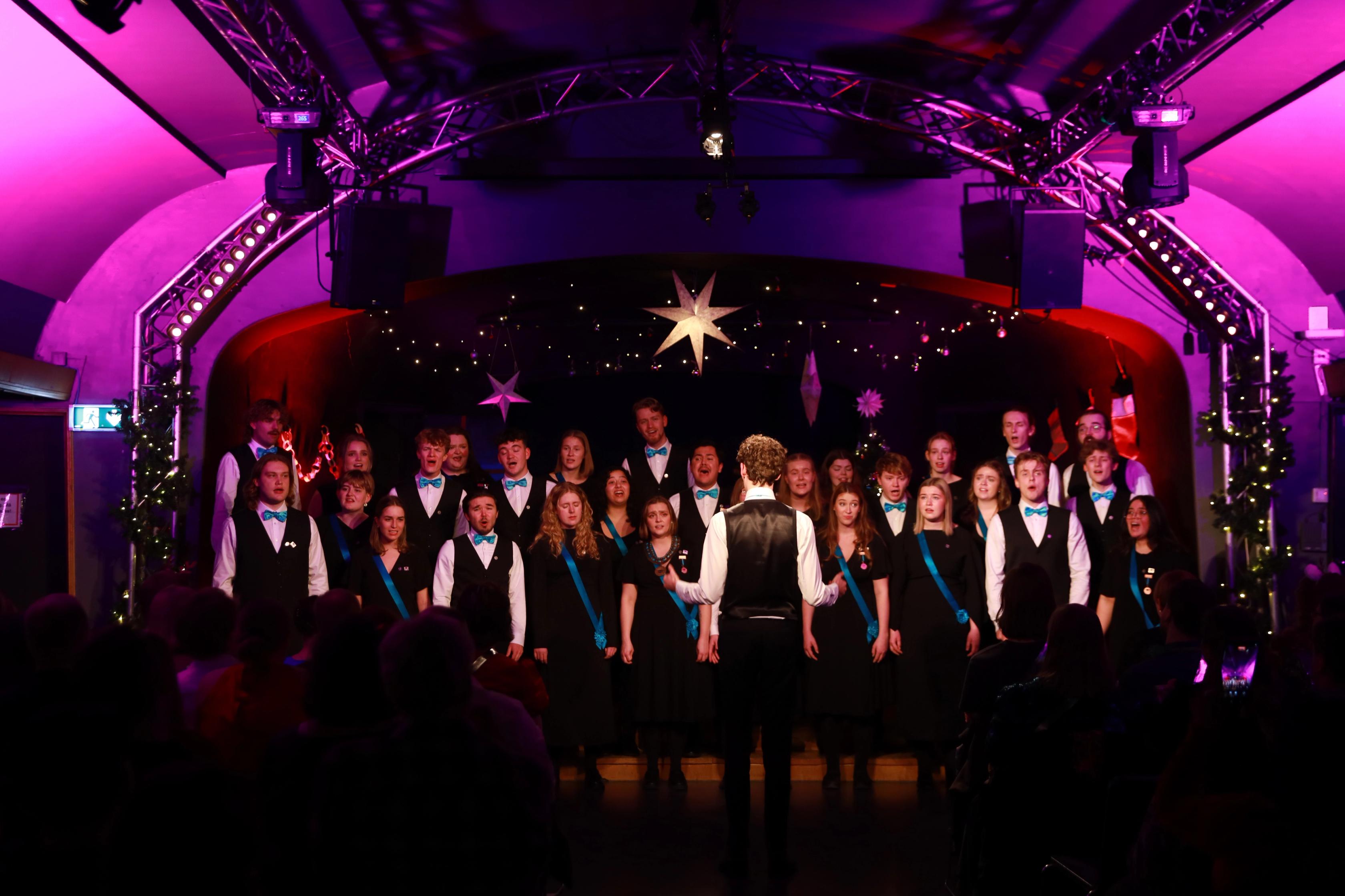 Et blandet kor i svarte antrekk med turkise detaljer synger på en julekonsert, ledet av en dirigent med ryggen til kameraet. Scenen er pyntet med lyslenker og store stjerner i taket, og publikum sitter i mørket foran.