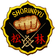 Et brodert Shorin Ryu-karateemblem med en gyllen knyttneve i midten, japanske tegn nederst og ordet «SHORINRYU» øverst på svart bakgrunn med gul kant.
