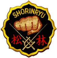 Et brodert Shorin Ryu-karateemblem med en gyllen knyttneve i midten, japanske tegn nederst og ordet «SHORINRYU» øverst på svart bakgrunn med gul kant.